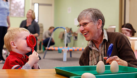 A young boy looking through a toy magnifying glass while the teacher looks on.